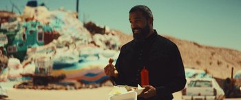Movie still from “Run & Gun” (2022), directed by Christopher Borrelli – A man with a beard is eating a hot dog; Medium shot, Over the shoulder angle