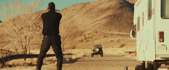 Movie still from “Run & Gun” (2022), directed by Christopher Borrelli – A man standing on the side of a road next to a jeep; Wide shot, Low angle