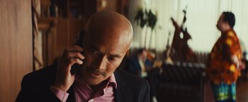 Movie still from “Run & Gun” (2022), directed by Christopher Borrelli – A bald man in a pink shirt talking on a cell phone; Close Up shot, Over the shoulder angle