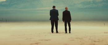 Movie still from “Run & Gun” (2022), directed by Christopher Borrelli – Two men in suits standing on the beach looking out at the ocean; Wide shot, Low angle