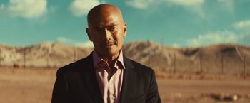 Movie still from “Run & Gun” (2022), directed by Christopher Borrelli – A bald man in a suit and pink shirt; Close Up shot, Low angle