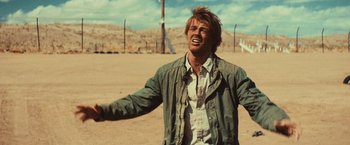 Movie still from “Run & Gun” (2022), directed by Christopher Borrelli – A man standing in the sand with his arms outstretched; Medium shot, Over the shoulder angle