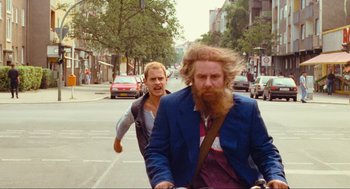 Movie still from “Run Lola Run” (1998), directed by Tom Tykwer – A man with a long beard riding a bike down a street; Medium shot, Over the shoulder angle