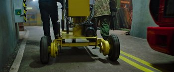 Movie still from “SAS: Red Notice” (2021), directed by Magnus Martens – Two men standing next to each other next to a yellow machine; Wide shot, High angle