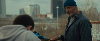 Movie still from “Samaritan” (2022), directed by Julius Avery – An older man in a blue hat is handing a box to a young boy; Medium shot, Over the shoulder angle