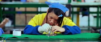 Movie still from “Samsara” (2011), directed by Ron Fricke – A woman in a yellow and blue jacket working on a piece of metal; Close Up shot, High angle