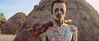 Movie still from “Samsara” (2011), directed by Ron Fricke – A person with a body painting holding a gun; Close Up shot, Low angle