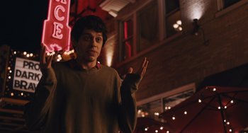 Movie still from “Big Time Adolescence” (2019), directed by Jason Orley – A young man is holding up a peace sign in front of a building; Medium shot, Over the shoulder angle