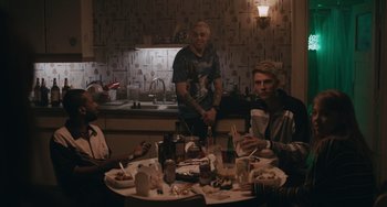 Movie still from “Big Time Adolescence” (2019), directed by Jason Orley – A group of people sitting at a table with plates of food on it; Medium shot, Over the shoulder angle