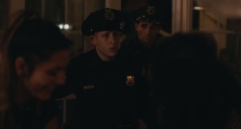 Movie still from “Big Time Adolescence” (2019), directed by Jason Orley – Two police officers are standing next to each other in a dark room; Medium shot, Over the shoulder angle