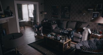 Movie still from “Big Time Adolescence” (2019), directed by Jason Orley – A man sitting on a couch in front of a coffee table; Wide shot, Over the shoulder angle