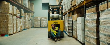 Movie still from “Savage Salvation” (2022), directed by Randall Emmett – A man sitting on the ground next to a forklift in a warehouse; Wide shot, High angle