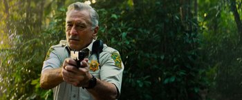 Movie still from “Savage Salvation” (2022), directed by Randall Emmett – An older man holding a video camera in front of trees; Close Up shot, Low angle