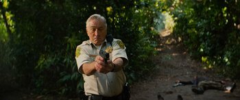 Movie still from “Savage Salvation” (2022), directed by Randall Emmett – An older man holding a gun while standing in front of trees; Medium shot, Low angle