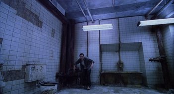 Movie still from “Saw” (2004), directed by James Wan – A man standing in a room under a light fixture; Wide shot, High angle