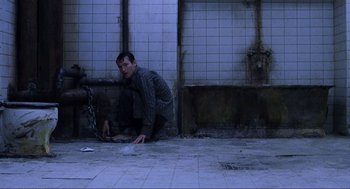 Movie still from “Saw” (2004), directed by James Wan – A man crouching on the ground with a chain around his neck; Wide shot, High angle