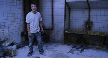 Movie still from “Saw” (2004), directed by James Wan – A young man standing in a bathroom with his bare feet in the floor; Wide shot, High angle