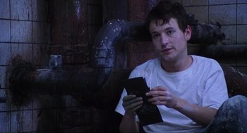 Movie still from “Saw” (2004), directed by James Wan – A young man holding a cell phone while sitting next to a pipe; Close Up shot, High angle