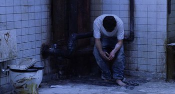 Movie still from “Saw” (2004), directed by James Wan – A man sitting on the ground in front of a pipe; Wide shot, High angle