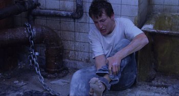 Movie still from “Saw” (2004), directed by James Wan – A man sitting on the ground with a pair of jeans on his feet; Medium shot, High angle