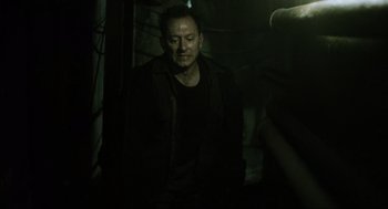 Movie still from “Saw” (2004), directed by James Wan – A man in a black jacket is sitting in a dimly lit room; Medium shot, Over the shoulder angle