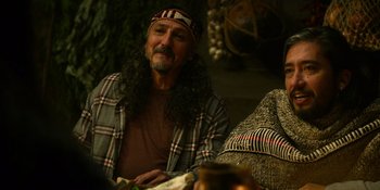 Movie still from “Sayen” (2023), directed by Alexander Witt – A man sitting on a couch wearing a plaid shirt; Medium shot, Over the shoulder angle