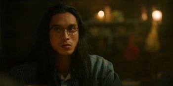 Movie still from “Sayen” (2023), directed by Alexander Witt – A man with long black hair wearing glasses and a jean jacket; Close Up shot, Over the shoulder angle