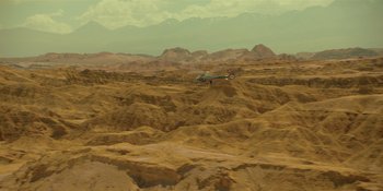 Movie still from “Sayen” (2023), directed by Alexander Witt – A helicopter flying over a desert landscape with mountains in the background; Extreme Wide shot, High angle