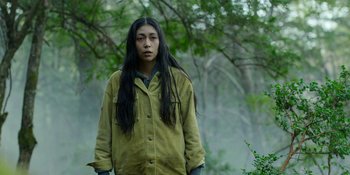 Movie still from “Sayen” (2023), directed by Alexander Witt – A person with long black hair standing next to a tree; Medium shot, Over the shoulder angle