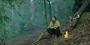 Movie still from “Sayen” (2023), directed by Alexander Witt – A person sitting on the ground near a fire in the woods; Wide shot, High angle