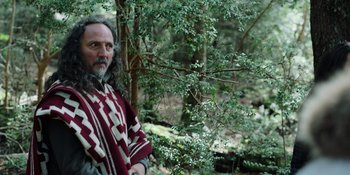 Movie still from “Sayen” (2023), directed by Alexander Witt – A man with a beard wearing a blanket standing in the woods; Medium shot, Over the shoulder angle