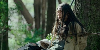 Movie still from “Sayen” (2023), directed by Alexander Witt – A woman with long hair sitting on a tree branch; Medium shot, High angle