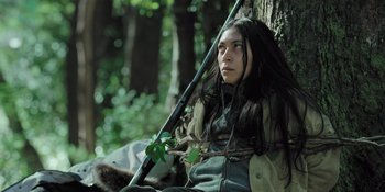 Movie still from “Sayen” (2023), directed by Alexander Witt – A person sitting on a tree branch holding a leaf; Medium shot, Low angle