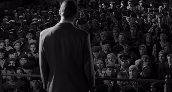 Movie still from “Schindler's List” (1993), directed by Steven Spielberg – A crowd of people sitting and standing in front of a man in a suit; Wide shot, High angle