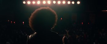 Movie still from “BlacKkKlansman” (2018), directed by Spike Lee – A person with a microphone in a dark room; Medium shot, Low angle