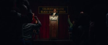 Movie still from “BlacKkKlansman” (2018), directed by Spike Lee – A man standing at a podium on a stage; Wide shot, High angle