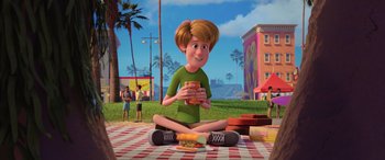 Movie still from “Scoob!” (2020), directed by Tony Cervone – A person sitting on the ground holding a sandwich; Wide shot, High angle