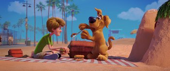 Movie still from “Scoob!” (2020), directed by Tony Cervone – A dog sitting on a blanket next to a person; Wide shot, High angle