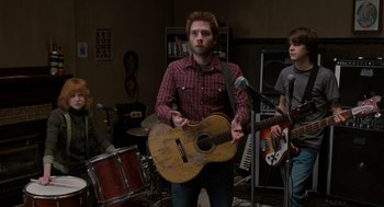 Movie still from “Scott Pilgrim vs. the World” (2010), directed by Edgar Wright – A man holding a guitar in front of two other men; Medium shot, Over the shoulder angle