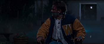 Movie still from “Scream” (1996), directed by Wes Craven – A man with a hockey jersey on sitting on a chair; Medium shot, Over the shoulder angle