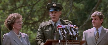 Movie still from “Scream 2” (1997), directed by Wes Craven – A man in a military uniform standing at a podium with microphones; Medium shot, Low angle