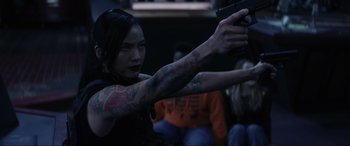 Movie still from “Secret Headquarters” (2022), directed by Henry Joost – A woman with tattoos holding a gun in front of a group of people; Close Up shot, Over the shoulder angle