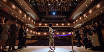 Movie still from “Secret Society of Second Born Royals” (2020), directed by Anna Mastro – A woman is cutting a purple ribbon in front of an audience; Extreme Wide shot, Low angle