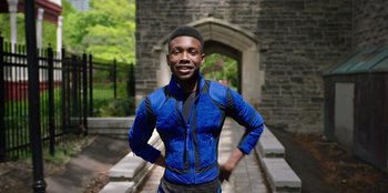 Movie still from “Secret Society of Second Born Royals” (2020), directed by Anna Mastro – A man in a blue jacket standing in front of a stone wall; Medium shot, Low angle