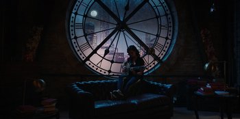 Movie still from “Secret Society of Second Born Royals” (2020), directed by Anna Mastro – A woman sitting on a couch in front of a large clock; Wide shot, High angle