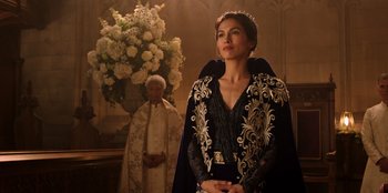 Movie still from “Secret Society of Second Born Royals” (2020), directed by Anna Mastro – A woman in a black and gold dress standing in front of an old man; Medium shot, Over the shoulder angle