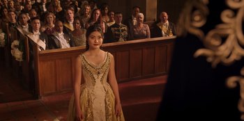 Movie still from “Secret Society of Second Born Royals” (2020), directed by Anna Mastro – A woman in a dress standing in front of a group of people; Medium shot, Over the shoulder angle