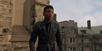 Movie still from “Secret Society of Second Born Royals” (2020), directed by Anna Mastro – A man in a suit standing in front of a building; Medium shot, Low angle
