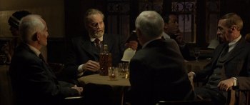 Movie still from “Blackthorn” (2011), directed by Mateo Gil – A man sitting at a table with a bottle of alcohol; Medium shot, Over the shoulder angle