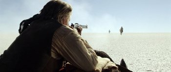 Movie still from “Blackthorn” (2011), directed by Mateo Gil – A man sitting on the side of a road holding a gun; Wide shot, Over the shoulder angle